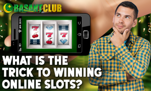 A man in a yellow and blue checkered shirt stands with his hand on his chin, looking thoughtful. A smartphone displaying an online slot machine with winning symbols (sevens, cherries, and diamonds) is shown. The bold text reads, "What is the trick to winning online slots?" The background features a green, celebratory theme with confetti.